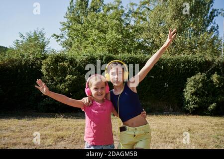 Le bambine abbracciano e ascoltano la musica con le cuffie gialle e rosa in giardino. Concetto estivo Foto Stock