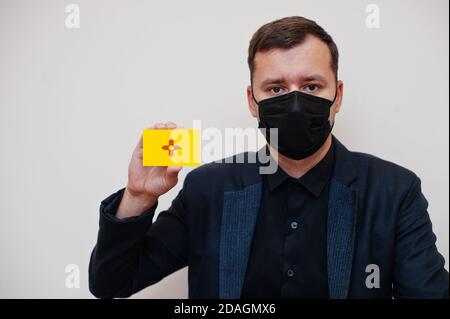 Uomo indossare nero formale e proteggere maschera facciale, tenere Alabama bandiera carta isolato su sfondo bianco. USA coronavirus Covid concetto paese. Foto Stock