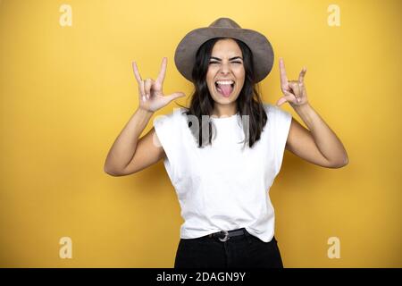 Bella donna che indossa una t-shirt bianca casual e un cappello in piedi su sfondo giallo gridando con espressione pazzesca facendo simbolo rock con le mani in su Foto Stock