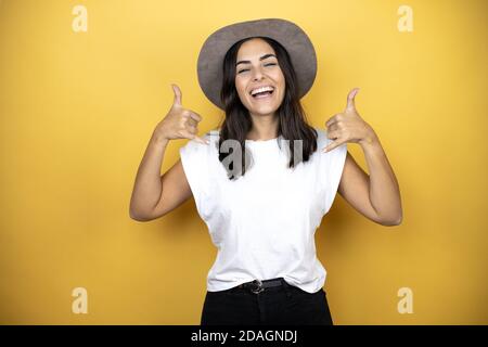 Bella donna che indossa una t-shirt bianca casual e un cappello in piedi su sfondo giallo gridando con espressione pazzesca facendo simbolo rock con le mani in su Foto Stock