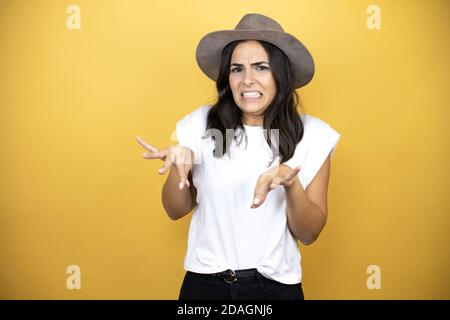 Bella donna che indossa casual t-shirt bianca e un cappello in piedi su sfondo giallo disgustava l'espressione, dispiaciuto e pauroso fare viso disgusto Foto Stock