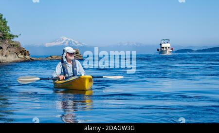 In una giornata estiva nel Mare di Salish, un uomo in un cappello da sole guarda dal suo kayak mentre un yacht a motore si avvicina, con Mt. La vetta innevata di Baker in lontananza. Foto Stock