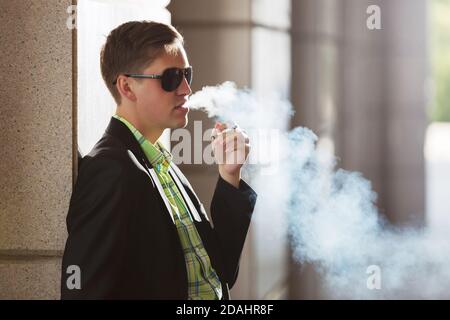 Giovane uomo in blazer nero e occhiali da sole che fuma una sigaretta sulla strada della città Foto Stock