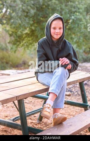 Tween Girl in una felpa con cappuccio. Ritratto di una ragazza teenage sorridente che indossa felpa con cappuccio verde, jeans e stivali gialli seduti sul tavolo di legno all'aperto. Foto Stock