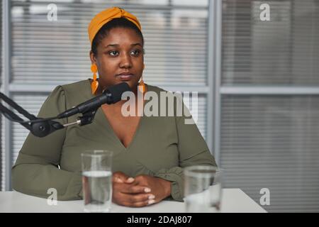 Donna africana sovrappeso che parla sul microfono mentre si siede a. il tavolo durante l'intervista alla radio Foto Stock