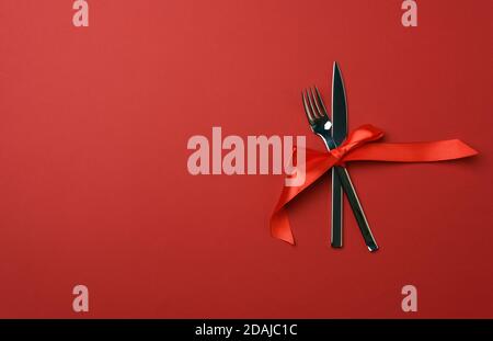 forchetta e coltello in metallo legato con un nastro di seta rossa, fondo rosso, posto per iscrizione, vista dall'alto Foto Stock
