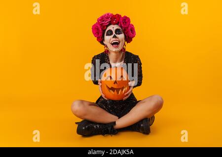 Ragazza giovane allegra nel trucco di Halloween con la zucca seduta sopra pavimento isolato su sfondo giallo Foto Stock