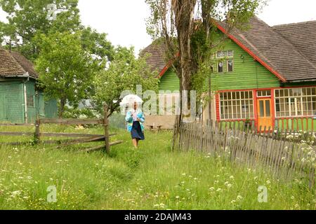 Contea di Brasov, Romania. Donna anziana che cammina attraverso il suo cortile. Tradizionali case in legno nei Monti Carpazi. Foto Stock