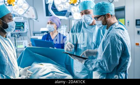 Chirurghi e assistenti professionali parlano e usano il computer Tablet digitale durante la chirurgia. Lavorano nella moderna sala operatoria dell'ospedale. Foto Stock