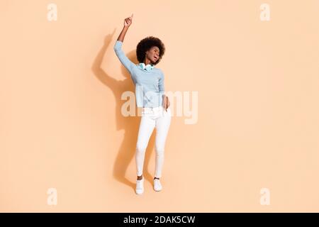 Foto ritratto a corpo intero di una donna spensierata e scura balla con le cuffie intorno al collo con un dito rivolto verso il basso isolato su beige pastello Foto Stock