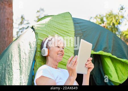 Donna anziana con cuffie e tablet PC davanti la tenda durante lo streaming o l'ascolto di musica Foto Stock