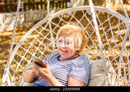 Donna anziana come pensionato nella poltrona da giardino legge Oppure scrive un SMS con lo smartphone Foto Stock