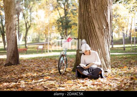 Giovane donna che legge un libro nel parco accanto alla sua bicicletta. È seduta sul terreno circondata da foglie cadute dagli alberi. Spazio per il testo. Foto Stock