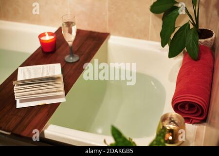 Tempo per te stesso. Rilassati a casa. Vasca da bagno con petali di fiori. Prenotate, candele e un bicchiere di vino su un vassoio di legno. Spa biologica relax nel comfort accogliente Foto Stock