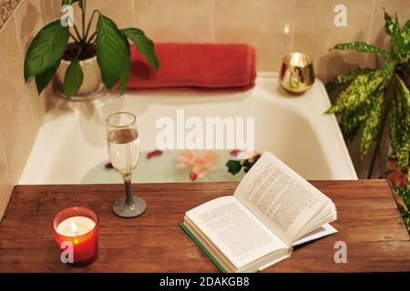 Tempo per te stesso. Rilassati a casa. Vasca da bagno con petali di fiori. Prenotate, candele e un bicchiere di vino su un vassoio di legno. Spa biologica relax nel comfort accogliente Foto Stock