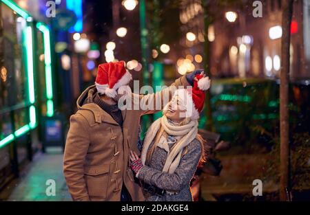 Una giovane coppia con Santa caps divertirsi in una passeggiata della città in una fredda notte. Relazione, insieme, Natale Foto Stock