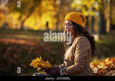 Ciao novembre. Donna trendy pensiva di 40 anni in cappotto beige e cappello arancione con foglie gialle d'autunno seduti all'aperto sul parco della città in autunno. Foto Stock