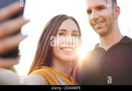 Felice coppia milleniale prendendo selfie all'aperto al tramonto - sorridente Amici che utilizzano lo smartphone in retroilluminazione - colori caldi del pomeriggio toni con messa a fuoco o Foto Stock