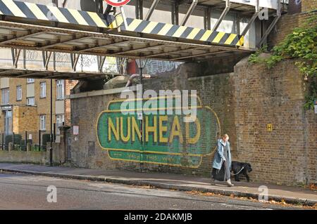 Una donna cammina il suo cane oltre il cartello dipinto Nunhead alla stazione Nunhead, a sud-est di Londra, Regno Unito. Sotto il ponte ferroviario su Gibbon Road. Foto Stock