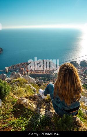 Foto verticale di una bruna ragazza seduta sulla cima della montagna srd guardando giù l'antica città croata di Dubrovnik. Caldo giorno estivo, vista del t Foto Stock