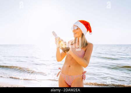 Bella donna bionda in bikini rosso e bianco e cappello Santa con champagne sulla spiaggia. Natale, nuovo anno in tropici. Vacanze, vacanze Foto Stock