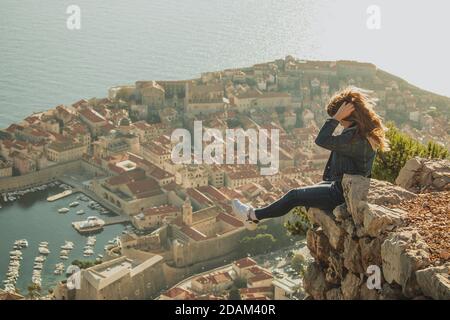 Felice eccitata ragazza seduta sul bordo di una roccia con la città vecchia di Dubrovnik dietro di lei. Bellissima città antica che risplende durante un tramonto pomeridiano, Foto Stock