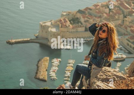 Bella brunetta vista da dietro in posa mentre si siede su una pietra sopra la città di Dubrovnik, contorni della maestosa città vecchia sullo sfondo. Foto Stock