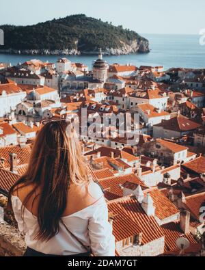 Vista posteriore verticale di una ragazza in piedi sulle mura della città di Dubrovnik, guardando verso i vecchi tetti rossi e le chiese in stretto impacchettato Foto Stock