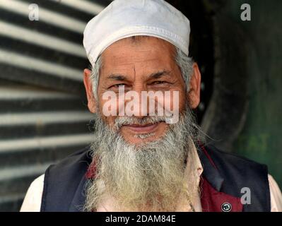 Anziano negoziante musulmano dell'India del Nord-Est con barba islamica e cappuccio di preghiera bianco pone e sorride per la telecamera. Foto Stock