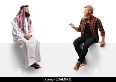 Hipster ragazzo bearded seduto su un pannello e che parla con un uomo arabo saudita in un tradizionale tobolo isolato sfondo bianco Foto Stock