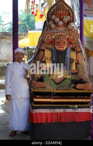 Il sacerdote balinese del tempio indù, in tradizionale abbigliamento bianco, si trova accanto a una maschera di Barong nei terreni di un tempio indù, nel centro di Bali, Indonesia Foto Stock