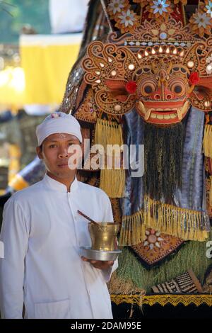 Il sacerdote balinese del tempio indù, in tradizionale abbigliamento bianco, si trova accanto a una maschera di Barong nei terreni di un tempio indù, nel centro di Bali, Indonesia Foto Stock