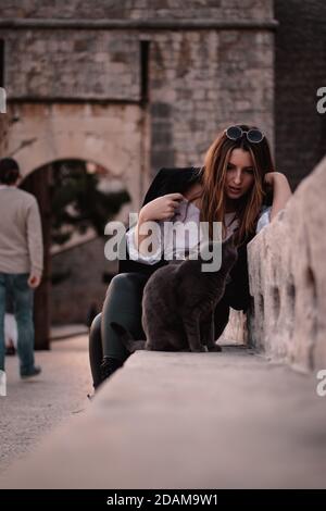 Donna seduta su un muro di pietra a dubrovnik, guardando un gatto grigio strada mentre la guarda indietro Foto Stock