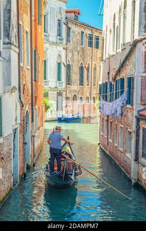 Venezia, Italia, 13 settembre 2019: Gondola a vela stretto canale tra vecchi edifici con mura di mattoni. Gondoliere vestito tradizionale bianco e blu a righe polo a maniche corte e cappello boater Foto Stock