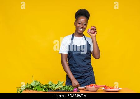 buona cuoca africana che tiene alcune cipolle con altri alimenti ingredienti e verdure davanti a lei su una scrivania Foto Stock