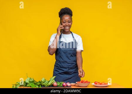 donna africana chef fare una telefonata, con ingredienti alimentari su un tavolo di fronte a lei Foto Stock