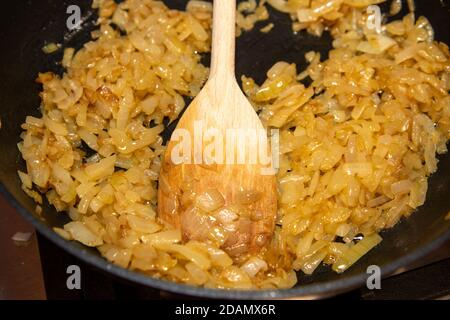 Le cipolle fritte cucinate in padella vengono spinte intorno con un cucchiaio di legno Foto Stock