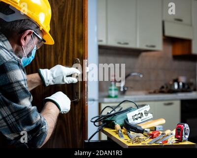 Falegname al lavoro di riparazione e installare una serratura della porta della stanza, indossare la maschera chirurgica per prevenire l'infezione da Coronavirus. Prevenire Pandemic Covid-19 a. Foto Stock