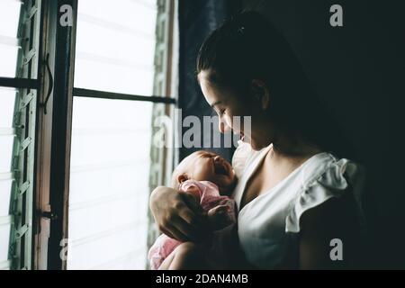 Una madre asiatica ha tenuto e guardato il bambino sorridente felicemente alla finestra tasto basso tono scuro. Foto Stock