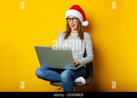 Giovane donna in un cappello di santa con un volto sorpreso si siede su una sedia e tiene un laptop ed è sorpreso a qualcosa su uno sfondo giallo. Banner. Foto Stock