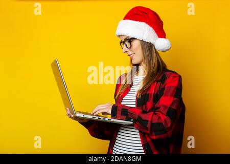 Giovane donna nel cappello di santa e camicia rossa in una gabbia tiene un laptop in mano e lavora su uno sfondo giallo. Foto Stock