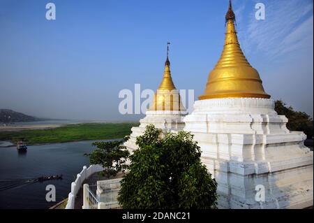 Una barca birmana passa due stupidi del tempio birmano con cima d'oro Sulle rive del fiume Ayeyarwady guardando oltre Sagaing vicino a Mandalay Myanmar Foto Stock