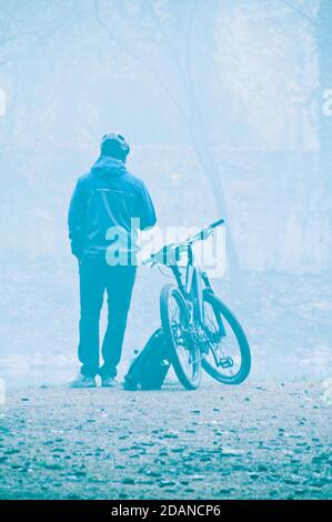 lone uomo in piedi vicino alla sua bicicletta di fronte ad un lago in una mattina foggy, riposante Foto Stock