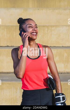 giovane e attraente donna africana che indossa abiti sportivi e tenuta guanti di pugilato mentre si prende per telefono e sorridendo su uno parcheggio Foto Stock