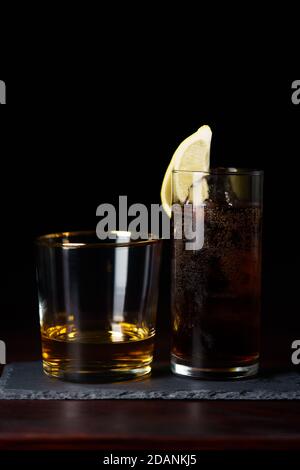 Whisky pulito e cola con ghiaccio e spicchio di limone servito su piastra di ardesia. Tavolo in legno rosso scuro, alta risoluzione Foto Stock