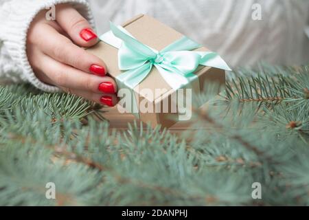 Una mano di donna in un maglione bianco lavorato a maglia con smalto rosso per unghie contiene un regalo con un nastro di menta, vicino ai rami di un albero di Natale. Foto Stock