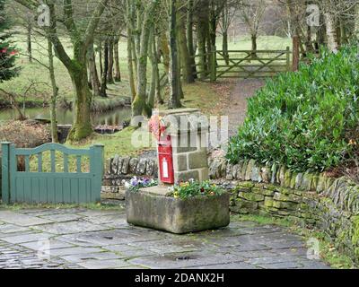 Piccolo postbox rosso posto in colonna di pietra su trogolo di pietra di fronte al muro di pietra bandiera pavimento cancello verde percorso acqua e alberi sullo sfondo Foto Stock