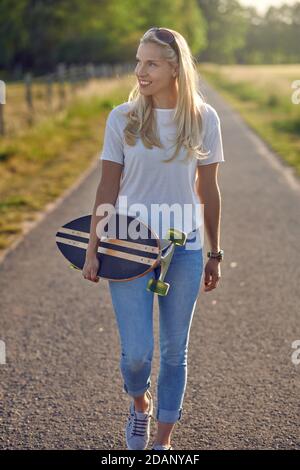 Si adatta giovane donna bionda sano che porta uno skateboard lungo un stretta strada di campagna guardando fuori dal lato con un felice sorrisi retroilluminati dal sole Foto Stock