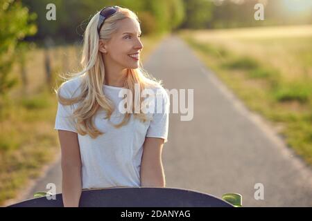 Si adatta giovane donna bionda sano che porta uno skateboard lungo un stretta strada di campagna guardando fuori dal lato con un felice sorrisi retroilluminati dal sole Foto Stock