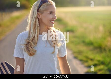 Si adatta giovane donna bionda sano che porta uno skateboard lungo un stretta strada di campagna guardando fuori dal lato con un felice sorrisi retroilluminati dal sole Foto Stock